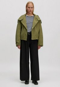 Olivenfarbene, übergroße Jacke mit Druckknopfverschlüssen und großem Kragen. Kombiniert mit einem gestreiften, taillierten Hemd und weiten schwarzen Hosen. Schwarze Schuhe.