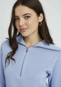 Pull en tricot bleu clair avec un col zippé et des manches longues, affichant un motif texturé et un détail de fermeture éclair en argent.