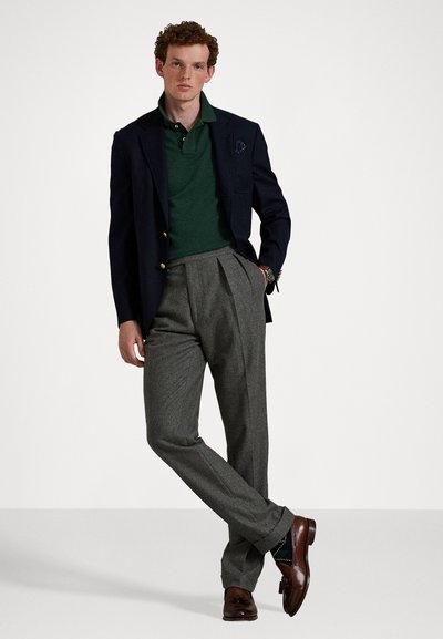 Blazer bleumarin cu butoane aurii, polo verde și pantaloni gri plisati. Mocasini maronii cu cusături decorative, pe un fundal alb.