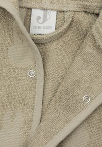 Beige badstof kledingstuk met een gestructureerd oppervlak, afgewerkt met een gladde, lichter gekleurde stofrand en drukknoopsluitingen. Maatlabel zichtbaar.