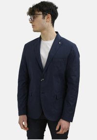 Blazer blu navy con un unico bottone, rever a punta, tasca frontale e tessuto textured. Indossato sopra una maglietta bianca.