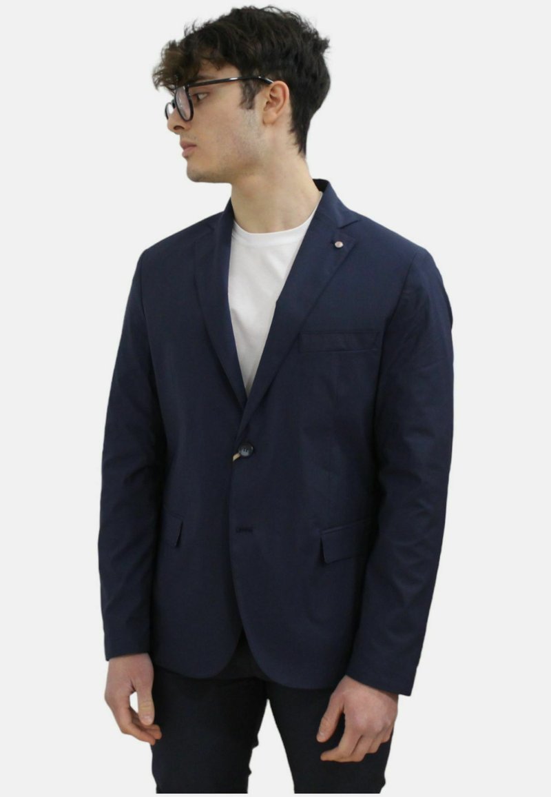 Blazer blu navy con un unico bottone, rever a punta, tasca frontale e tessuto textured. Indossato sopra una maglietta bianca.