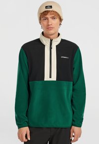 Groen en zwarte kwart-rits pullover van zacht materiaal, met een crèmekleurig paneel en een logo op de borst. De beanie is beige.