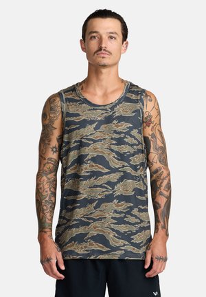 Homme aux cheveux lissés en arrière et tatouages sur les bras, portant un débardeur camouflage sans manches et un short noir, sur un fond uni.
