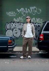 Béžová károvaná košeľa, biele polo s textom, hnedé nohavice s tenkými pásikmi, biele tenisky. Mestské prostredie s autami a pozadím s graffiti.