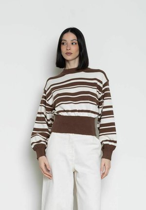 Donna con capelli neri e lisci che indossa un maglione a righe marroni e bianche e pantaloni bianchi, in piedi davanti a uno sfondo chiaro e uniforme.