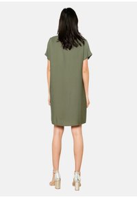 Oliwkowa sukienka krótko rękawowa o luźnym kroju, do kolan, z teksturowanej tkaniny oraz okrągłym dekoltem. Połączona ze srebrnymi sandałami na obcasie.