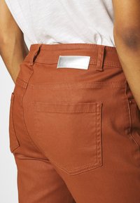 Rust-colored pants made of smooth fabric feature a silver logo patch on the waistband, two back pockets, and tailored stitching.