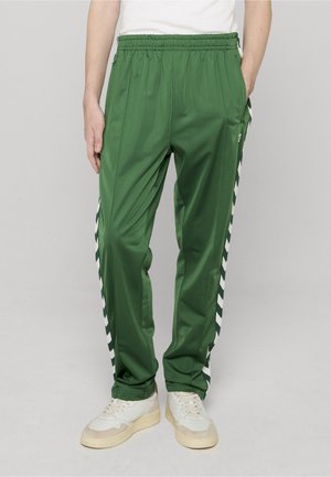 Groene trainingsbroek gemaakt van een glad, glanzend materiaal met witte chevronstrepen langs de zijkanten en een elastische tailleband.