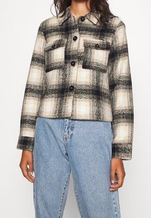 Vrouw draagt een zwart-beige-wit geruite jas met knopen en twee voorzakken, gecombineerd met lichtblauwe hooggetailleerde jeans.