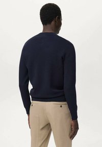 Suéter de punto azul marino con cuello redondo y dobladillo acanalado, combinado con pantalones beige claros que presentan dobles bolsillos traseros y detalles de botones.