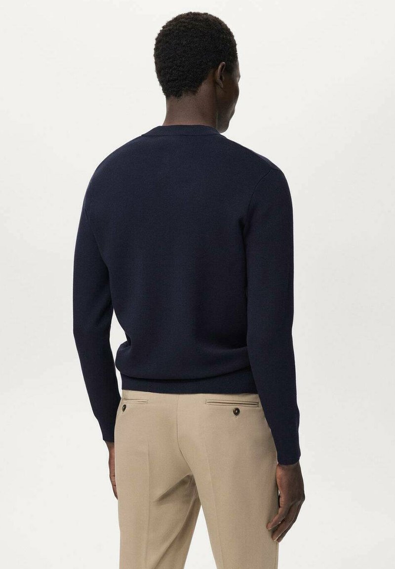 Suéter de punto azul marino con cuello redondo y dobladillo acanalado, combinado con pantalones beige claros que presentan dobles bolsillos traseros y detalles de botones.