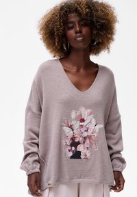 Vrouw met krullend haar, gekleed in een losse mauve trui met een artistieke print van een vrouwen gezicht bedekt met roze bloemen en vlinders.