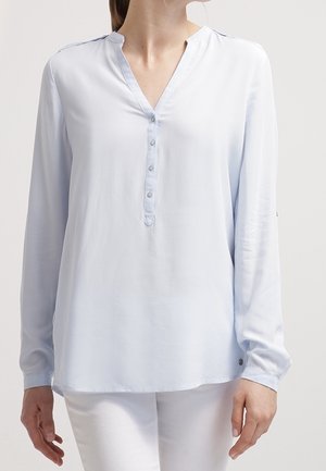 Kvinde iført en lyseblå bluse med lange ærmer, V-hals og knappet front, kombineret med hvide bukser, foran en ensfarvet baggrund.