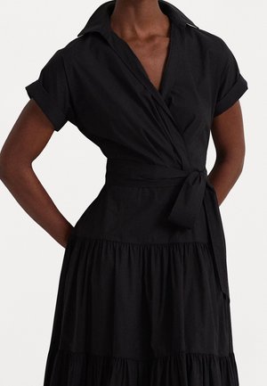 Nainen, jolla on lyhythihainen musta kietaisumekko, jossa on käärityt kalvosimet ja vyötä, seisomassa käsivarret lanteilla yksiväristä taustaa vasten.