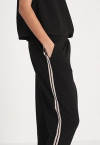 Svarta byxor med avslappnad passform, med sidfickor och vertikala beige ränder längs varje ben. Tillverkade av ett mjukt, lätt tyg.