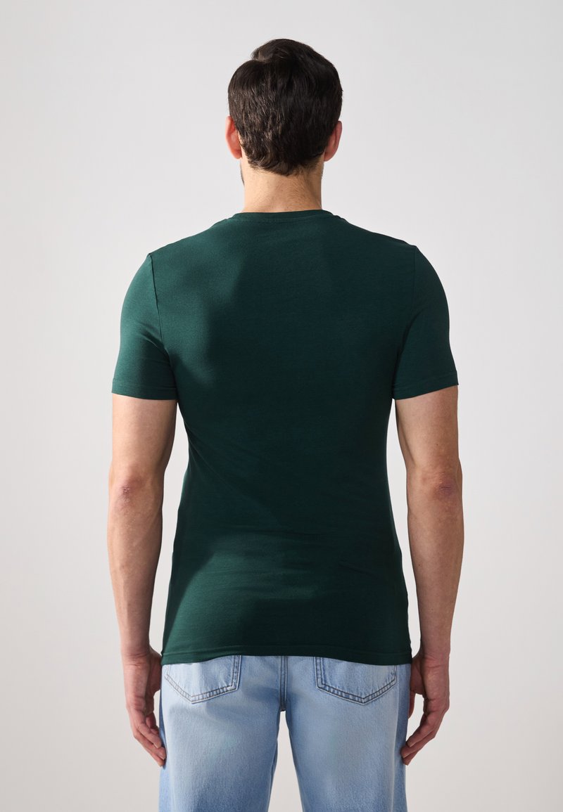 Ciemnozielona koszulka z krótkim rękawem, dopasowany krój, gładka faktura, okrągły dekolt, warstwowy dół. Noszona z jasnoniebieskimi dżinsami, widoczne detale szycia.