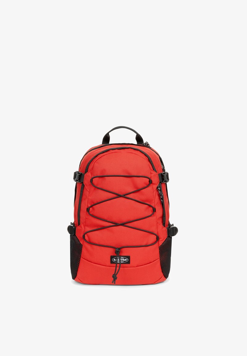 Zaino in nylon rosso con tasche laterali in rete nera, spallacci regolabili e dettagli in corda elastica sul davanti. Scomparti con zip per la conservazione.