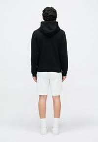 Svart hettegenser med stor hette, kombinert med hvite shorts. Modellen står i et nøytralt miljø og viser ryggen.