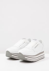 Baskets plateforme blanches avec une tige en cuir lisse, des détails perforés, une semelle épaisse à rayures grises, et des lacets blancs plats. Les accents incluent un talon gris.