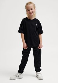 Schwarzes, oversized T-Shirt mit kleinem, weißem Logo auf der Brust, kombiniert mit schwarzen Hosen und weißen Sneakers mit grauen Akzenten.