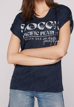 Frau trägt ein dunkelblaues kurzärmeliges Hemd mit hochgekrempelten Ärmeln und weißem Text, kombiniert mit hellblauen Jeansshorts.