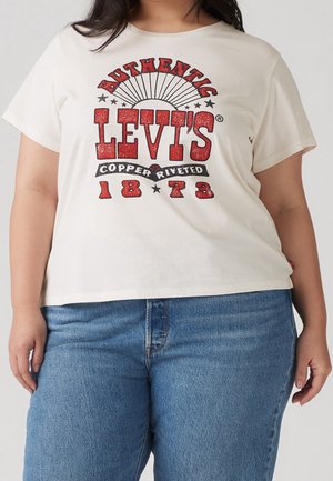 Žena oblečená v bílém tričku Levi's s červeným a černým textem "Autentické Levi's s měděnými nýtky 1873" a modrých džínách.