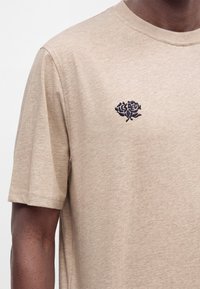 Μπεζ κοντομάνικο T-shirt με μία μικρή μαύρη κεντημένη τριαντάφυλλα και κείμενο στο αριστερό στήθος, που φοριέται από άτομο με σκούρο χρώμα δέρματος.
