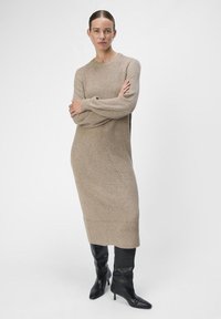 Beige gebreide sweaterjurk met lange mouwen, ribbelstructuur en zijdetails, gecombineerd met hoge zwarte puntige laarzen.