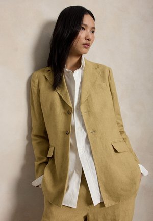 Γυναίκα με μακριά σκούρα μαλλιά, ντυμένη με σακάκι και παντελόνι μουσταρδί χρώματος πάνω από λευκό πουκάμισο με κουμπιά, στέκεται μπροστά σε έναν απλό μπεζ τοίχο.