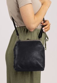 Black leather crossbody bag with a textured finish, silver hardware, and a simple design, worn at the side against a green outfit.