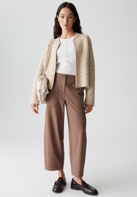 Beige flauschige Jacke über einem weißen Oberteil, kombiniert mit hochgeschnittenen braunen weiten Hosen und schwarzen Loafers. Inklusive einer beige Schultertasche.