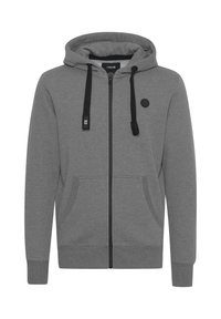 Sudadera con cremallera gris hecha de una tela suave. Presenta una capucha con cordón, detalles en negro, dos bolsillos frontales y un logo redondo en el pecho.