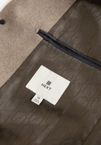 Interno di giacca marrone chiaro con fodera marrone strutturata, tasca con zip e un'etichetta di stoffa bianca con il logo "NEXT" e il tag della taglia M.