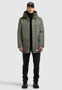 Olivengrøn polstret parka med hætte, lynlås foran, sidelommer og sorte detaljer, kombineret med sorte jeans og sorte sko.