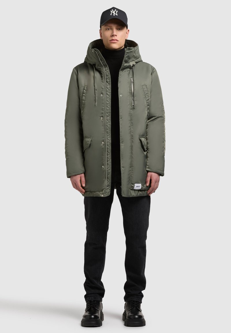 Olivengrøn polstret parka med hætte, lynlås foran, sidelommer og sorte detaljer, kombineret med sorte jeans og sorte sko.