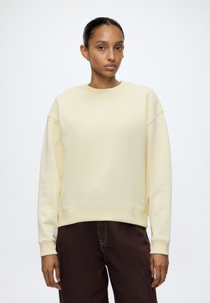 Femme portant un sweat-shirt jaune clair uni à manches longues et un pantalon marron foncé, debout devant un fond blanc.
