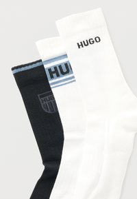 Τρία ζευγάρια κάλτσες: ένα μαύρο με μπλε λεπτομέρειες, ένα λευκό με μπλε ρίγες και ένα μονόχρωμο λευκό, όλα με το λογότυπο "HUGO".