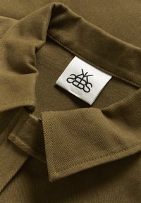 Olivgrüner Cordstoff mit strukturiertem Oberfläche, ausgestattet mit einem umgeschlagenen Kragen und einem weißen Etikett mit schwarzem Logo.