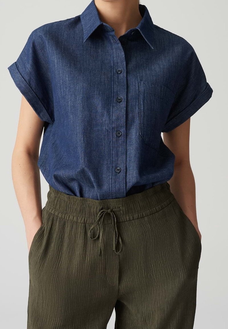 Femme portant une chemise à manches courtes bleu foncé boutonnée, rentrée dans un pantalon kaki texturé à taille élastique, mains dans les poches.