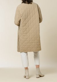 Quilted beige long coat with side slits, made from lightweight material. Paired with white cropped jeans and beige ankle boots.
