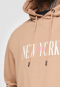 Felpa con cappuccio color beige che presenta una grafica frontale con la scritta "NEW YORK" in bianco con una sottolineatura rosa, coulisse con punte nere e una texture morbida.