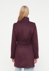 Violets mētelis ar augstu apkakli, jostotu vidukli un garām piedurknēm; teksturēta auduma, ar gludu apdari un taisnu siluetu.