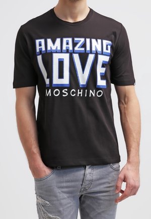 Mies pukeutuneena mustaan Moschinon T-paitaan, jossa on sinivalkoinen teksti "AMAZING LOVE", yhdistettynä vaaleanharmaisiin kulutettuihin farkkuihin.