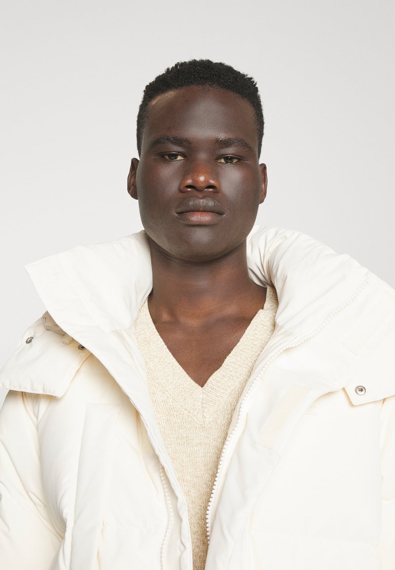 Doudoune blanche avec col haut, tissu texturé et coutures visibles, portée sur un pull beige à col en V. Design minimaliste.