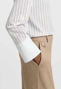 Person in einem beigen, gestreiften Langarmhemd mit weißen Manschetten und beigen Hosen, mit einer Hand in der Tasche.