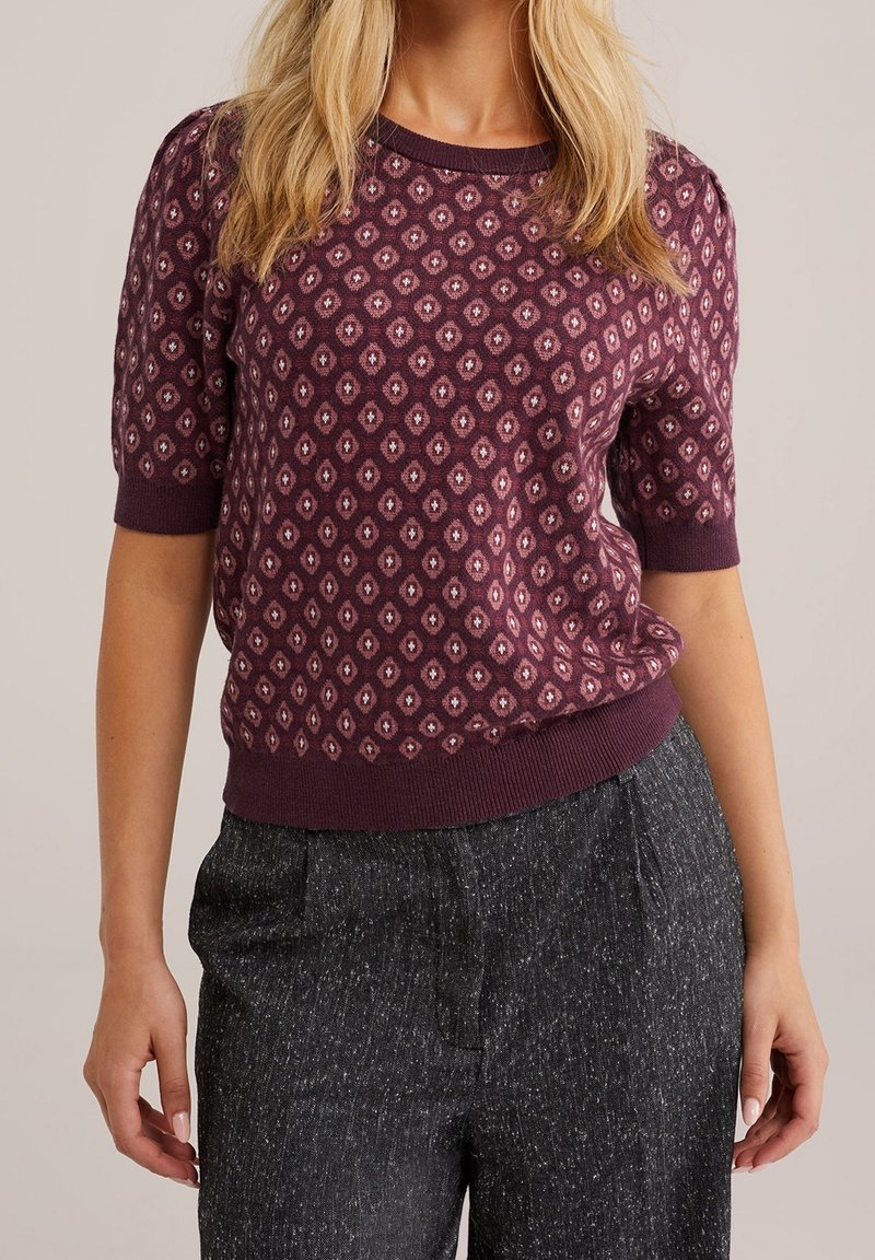 Femme portant un pull violet à manches courtes à motifs et un pantalon taille haute gris foncé texturé sur un fond uni.