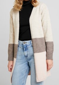 Beige, brun och ljusrosa färgblockerad cardigan med ribbade ärmslut, öppen frontdesign och mjukt stickat tyg. Bärs över en svart topp.