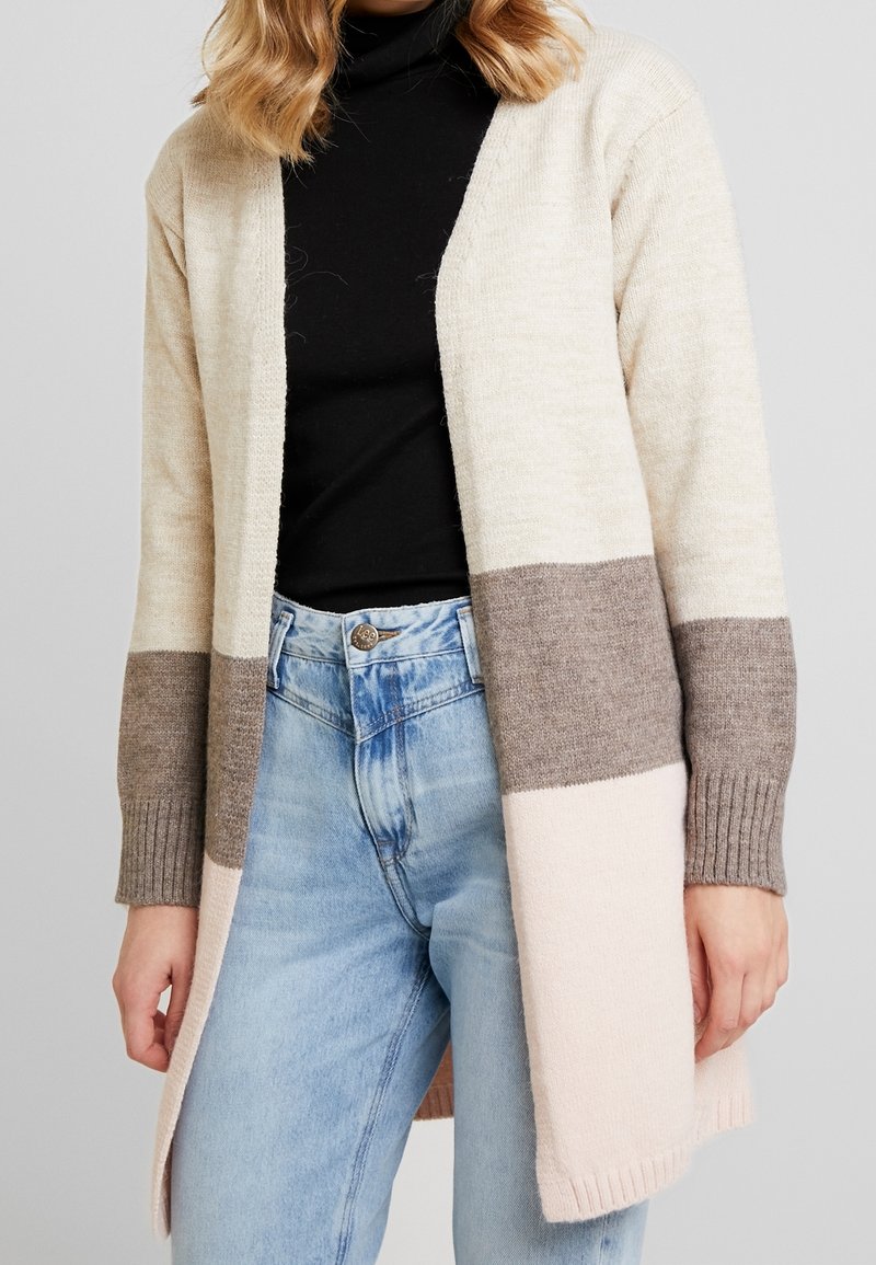 Beige, brun och ljusrosa färgblockerad cardigan med ribbade ärmslut, öppen frontdesign och mjukt stickat tyg. Bärs över en svart topp.