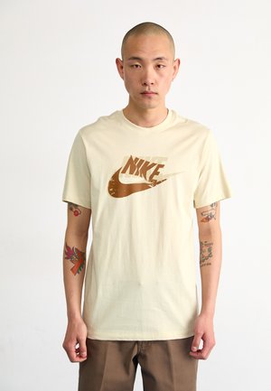 T-shirt corto a maniche corte color crema con il logo Nike marrone sul petto. Materiale in cotone, vestibilità comoda, texture morbida. Senza fantasie o dettagli.